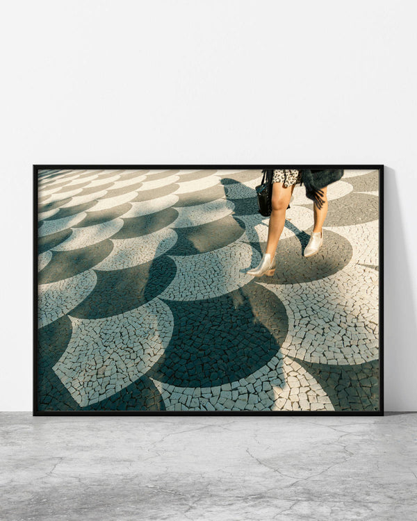Geometric print of a person walking across sunlit hand-laid scallop-pattern stone pavement with a long shadow