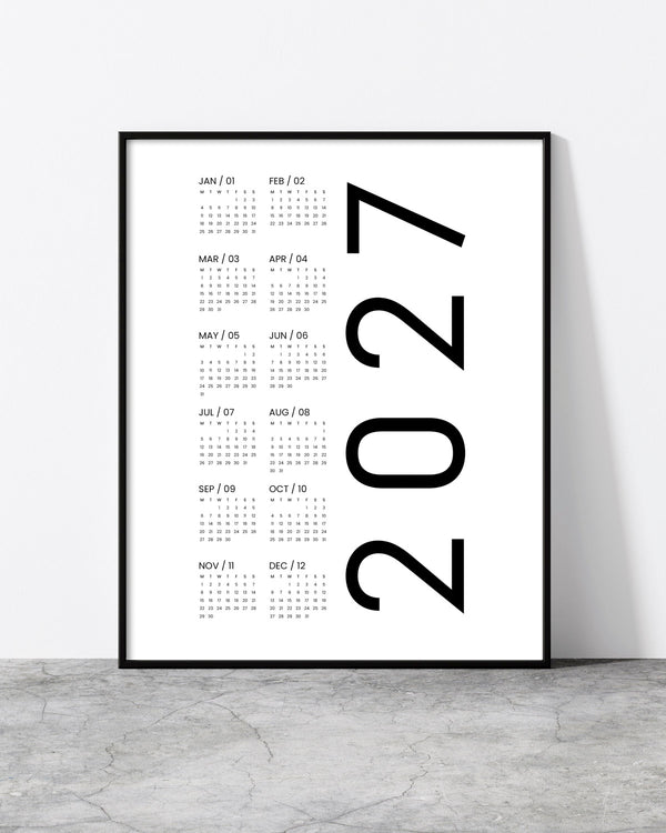 A minimalist framed 2027 calendar print leaning against a white wall on a gray concrete floor. The print features a clean white background with the years '2027' in large black font next to a vertical list of monthly calendar grids in small gray text, enclosed in a thin black frame.