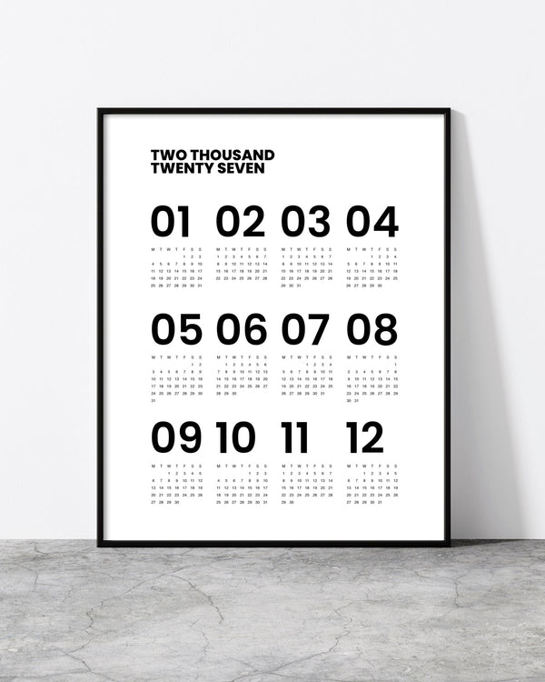 A minimalist framed calendar print leaning against a white wall on a gray concrete floor. The print features the text 'TWO THOUSAND TWENTY SEVEN' at the top and the numbers 01 through 12 with small dot-based numerals underneath, all in black on a white background.