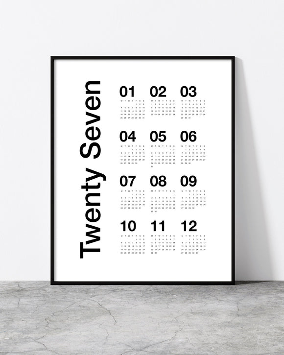 A minimalist black-framed wall calendar print leaning against a white wall on a gray concrete floor. The print features the text 'Twenty Seven' vertically aligned next to a grid of numbers from 01 to 12, each with small dotted calendar blocks underneath.