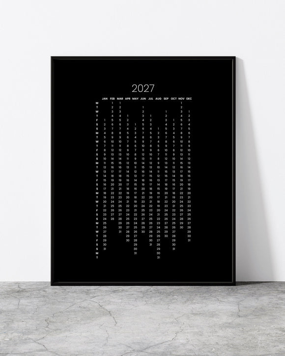 A black framed vertical wall art print featuring the year 2027 in white text, leaning against a white wall on a light gray stone floor.