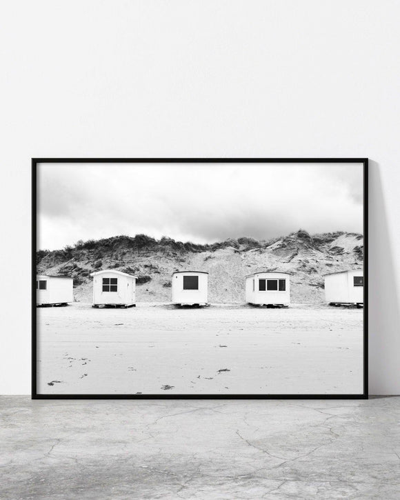Monochrome image of small beach huts standing in a row at the base of rugged sand dunes.