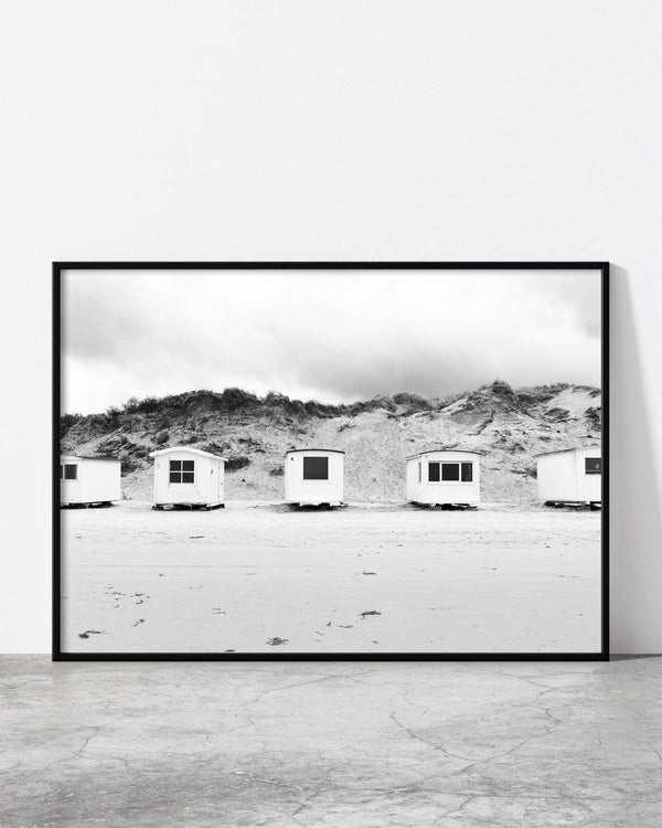 Monochrome image of small beach huts standing in a row at the base of rugged sand dunes.