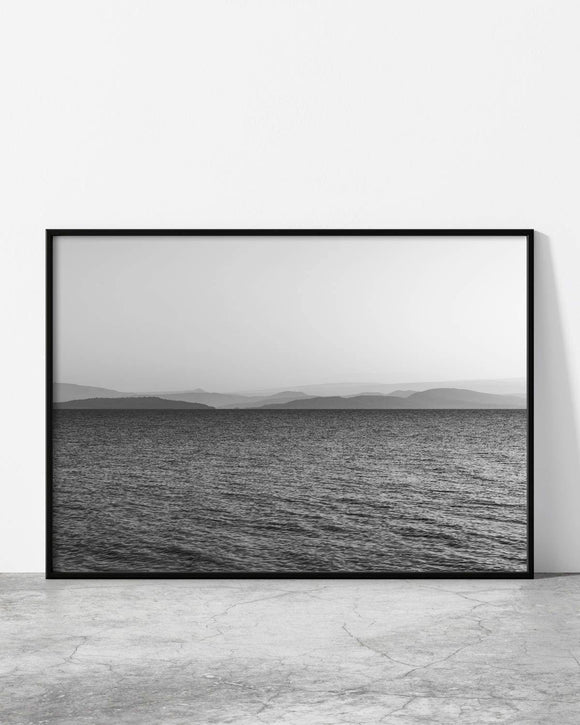 Black and white landscape photograph of a calm sea with distant mountain silhouettes under a clear sky.