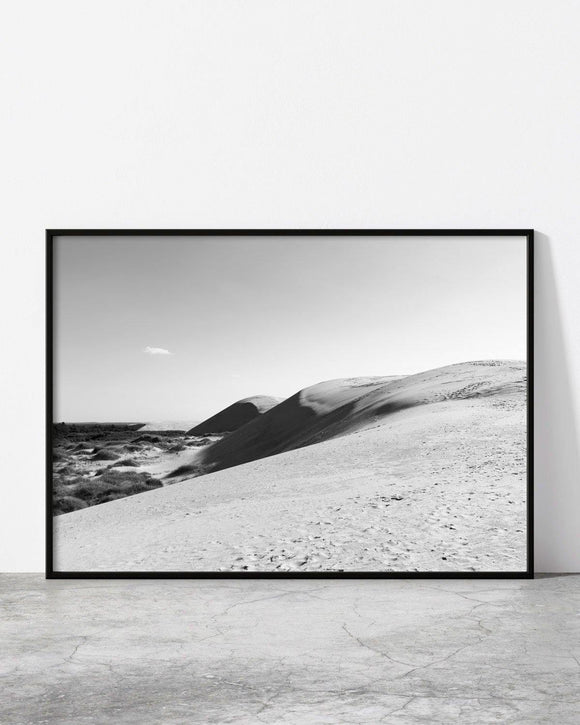 Black and white photo of rolling sand dunes with footprints leading into the distance under a clear sky.