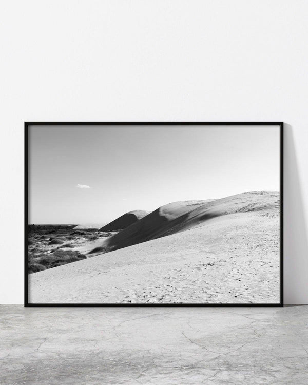 Black and white photo of rolling sand dunes with footprints leading into the distance under a clear sky.
